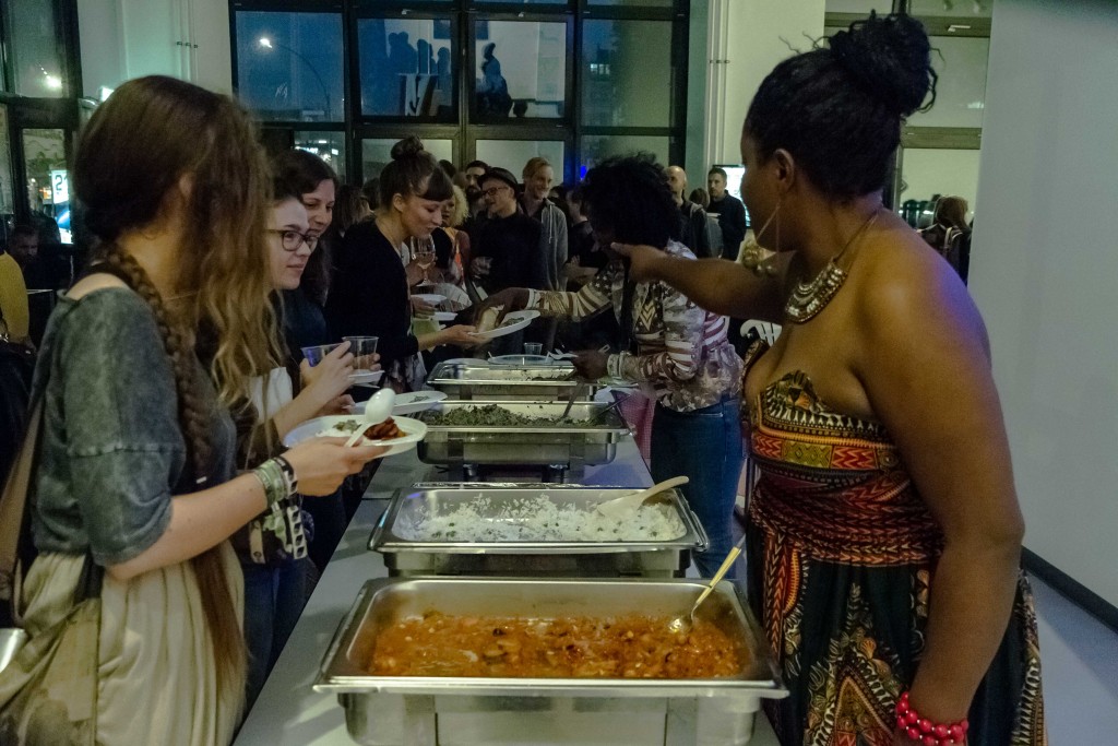 Die Ausstellungsbesucher stellen sich vor einem Buffet mit afrikanischen Speisen auf, welche ihnen durch zwei junge Frauen gereicht werden. Eine davon trägt ein Kleid im afrikanischen Stil.