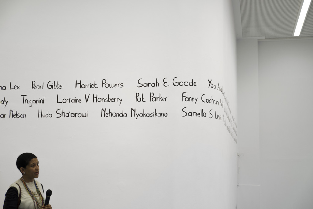 Die Künstlerin steht vor der Wand mit den aufgemalten Frauennamen und führt Erläuterungen aus, ein Mikrofon in der Hand haltend.