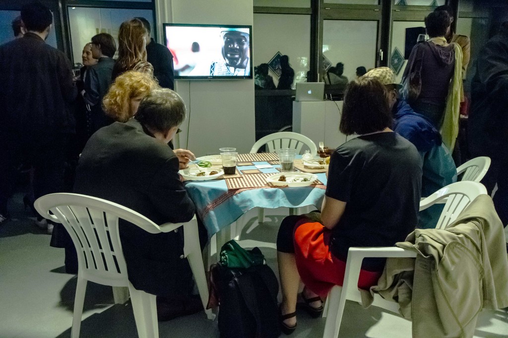 Eine kleine Gruppe von Besuchern isst beim Tisch, welcher zusammen mit mehreren Stühlen zu den ausgestellten Kunstwerken gehört, und schaut einen Film, der ebenfalls einen Teil der Ausstellung bildet.