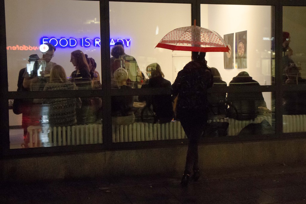 Eine Frau steht unter einem Regenschirm vor der verglasten Galeriefront und schaut ins Innere zu den sich im regen Austausch befindenden Besuchern.